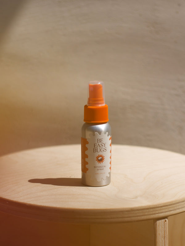 Be Easy Bugs Spray in a 2oz aluminum bottle with orange plastic cap sitting on a wooden stool in front of a mud colored wall